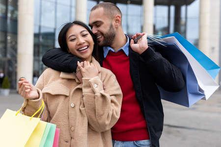 Portrait of happy multiracial couple with shopping bags smiling and hugging near supermarketの写真素材