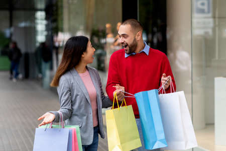 Excited international couple with gift bags standing near shopping mall, shouting OMG, overjoyed about seasonal salesの写真素材