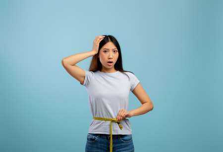 Sad and disappointed asian lady measuring waist with tape, unhappy about her body parameters, blue backgroundの写真素材
