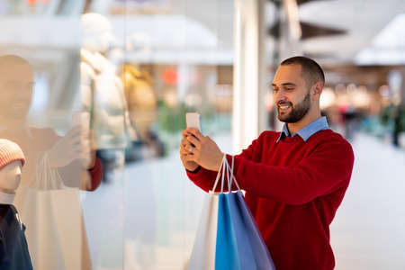 Happy millennial guy with shopper bags using smartphone at supermarket, taking selfie or photo, chatting with friendの写真素材