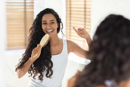 Cheerful Young Woman Singing With Hairbrush While Looking In Mirror In Bathroom