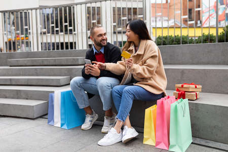 Cheerful interracial couple sitting on stairs outdoor, using mobile phone and credit card, surrounded with shopping bagsの写真素材