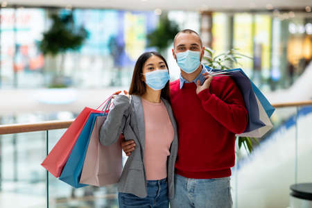 Portrait of young multiracial couple with shopper bags wearing face masks, looking at camera in modern supermarketの写真素材