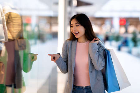 Gorgeous young Asian woman with shopping bags smiling and pointing at store window in supermarketの写真素材