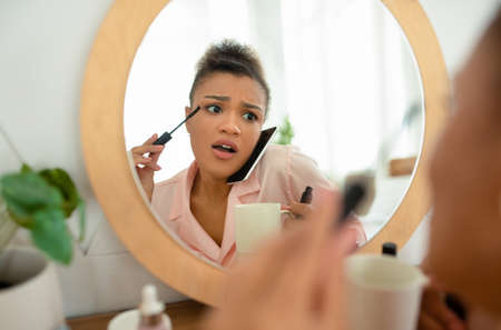 Emotional african american lady talking on cellphone with girlfriend while putting mascara in front of mirrorの写真素材