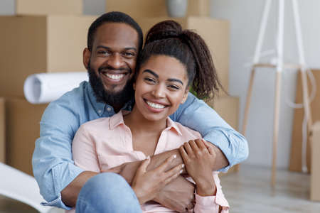 Satisfied young african american wife and husband hugging in new apartment with boxesの写真素材