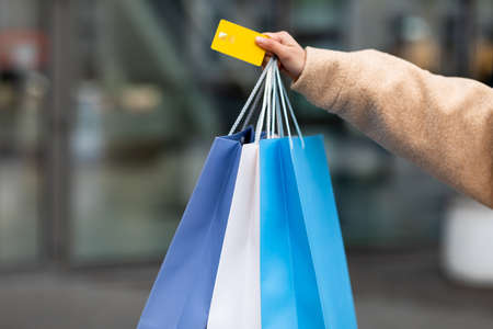 Unrecognizable young woman in winter coat holding credit card and shopper bags outdoors, shopping for cold seasonの写真素材