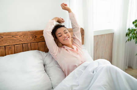 Good start of new day. Well-slept young black lady stretching arms after waking up, sitting in bed in cozy bedroomの写真素材