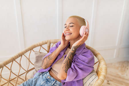 Calm young woman listening to music, audio book, podcast in wireless headphones, sitting in wicker chair, copy spaceの写真素材