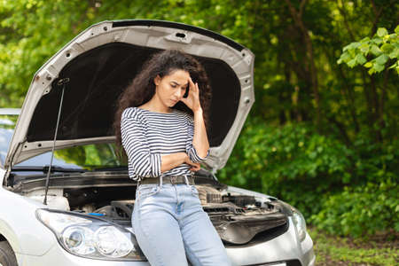 Puzzled woman driver leaning on open car hood