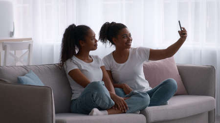 Cheerful young black sisters making selfie with smartphone at homeの写真素材
