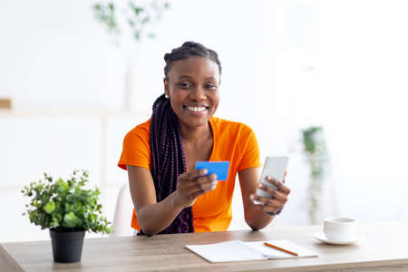 Young African American woman buying online with credit card and mobile phone, sitting at desk...