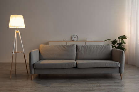 Sofa, clock, potted plant and glowing lamp in evening on gray wall background in office or living roomの写真素材