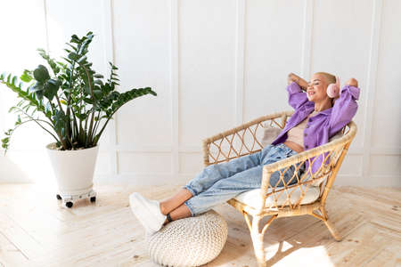 Full length of relaxed lady listening to music in wireless headphones, resting in wicker armchair in light roomの写真素材