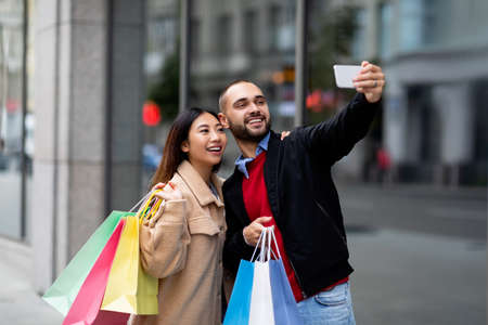 Cheerful multiracial couple with shopper bags taking selfie on smartphone in front of big supermarketの写真素材