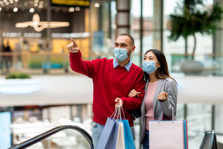 Millennial diverse couple wearing protective masks while shopping at supermarket, holding shopper bags, copy space
