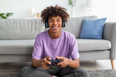 Portrait of cool black teen gamer with controller playing online video games, wearing headphones at homeの写真素材