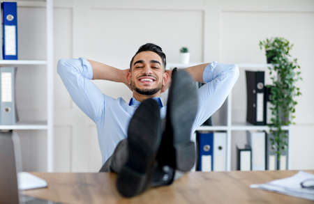 Happy millennial Arab man relaxing after successful work, putting feet on table at modern officeの写真素材