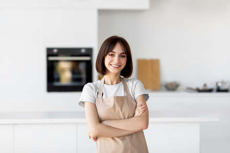 Young caucasian lady in apron standing with folded arms, looking and smiling at camera, posing in...
