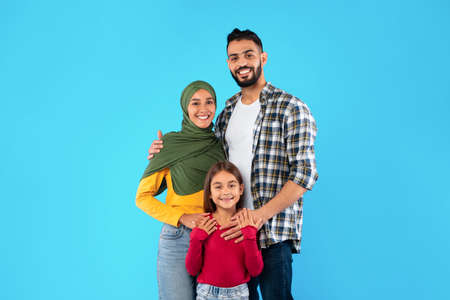 Modern Muslim Family Of Three Hugging Standing Over Blue Backgroundの写真素材