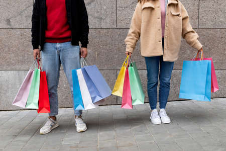Unrecognizable young couple holding shopper bags, posing in front of stone wall outdoors, going shopping togetherの写真素材