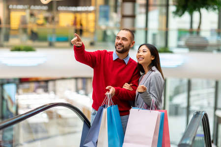 Happy diverse couple with gift bags using moving stairway, going shopping together at city mallの写真素材