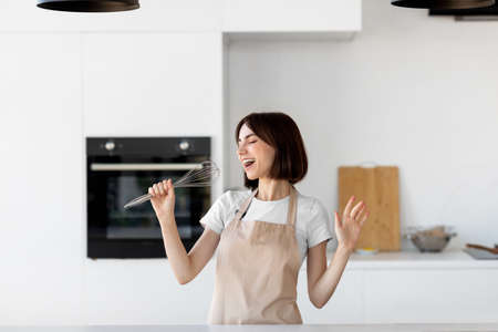 Young happy housewife dancing and singing a song in modern kitchen interior, pretending whisk to be a microphoneの写真素材