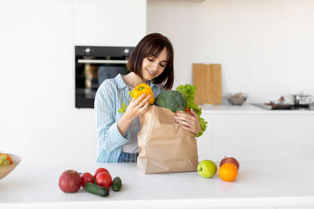 Millennial woman unpacking paper bag with fresh organic vegetables and fruits, standing in modern kitchenの写真素材