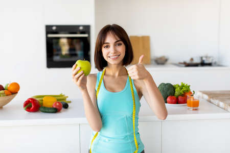 Young slim woman holding apple and showing thumb up, recommending healthy eating, posing in kitchen...