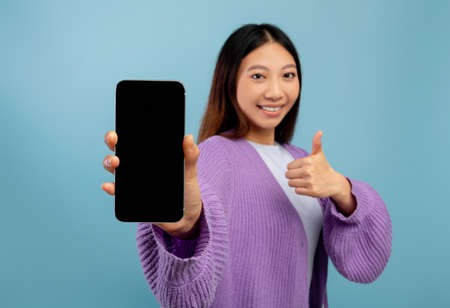 I recommend. Happy asian lady showing smartphone with blank screen, gesturing thumb up and smiling, blue backgroundの写真素材