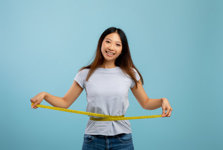 Weight loss concept. Fit asian lady measuring her waist with tape over blue studio backgroundの写真素材
