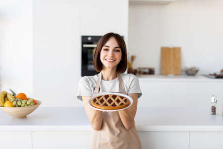 Homemade dessert. Happy lady holding and showing freshly baked pie, wearing apron and preparing pastry in kitchenの写真素材