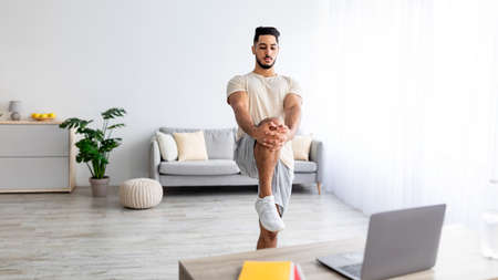 Young Arab man stretching his leg in front of laptop during domestic training, following online video tutorial on laptopの写真素材