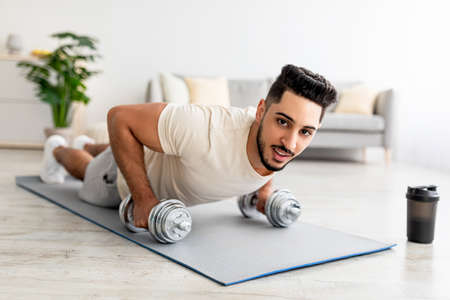 Stay at home fitness. Strong young Arab man doing whole body workout, standing in plank pose, using dumbbells indoorsの写真素材