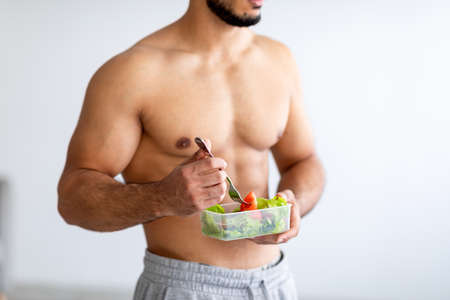 Balanced diet concept. Cropped view of strong young man with bare torso eating vegetable salad at homeの写真素材