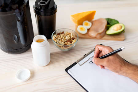 Closeup of young bodybuilder writing meal plan on clipboard with mockup, eating healthy foods, using protein shakesの写真素材