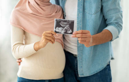 First portrait of our baby. Happy muslim pregnant woman and her husband showing sonogram picture to cameraの写真素材