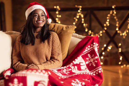 Cozy Christmas atmosphere. Young black woman in Santa hat sitting on couch under warm plaid, celebrating Xmas at homeの写真素材