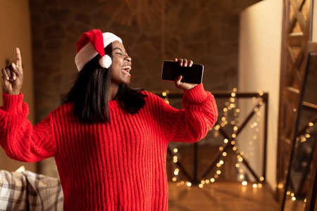 Christmas holiday music. Happy black lady in Santa hat singing, using smartphone as mic at home, mockup for mobile appの写真素材