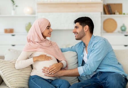 Happy muslim couple expecting baby, resting on couch at home and smiling, man touching his pregnant wifes bellyの写真素材