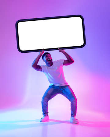 Black man holding big smartphone with empty screen in neon light. Mockup for advertisement, mobile...