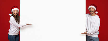 Christmas Ad. Cheerful Couple In Santa Hats Pointing At White Advertisement Boardの写真素材