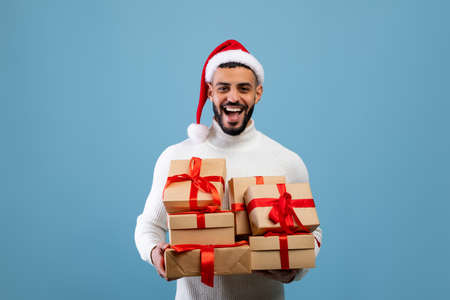 Buying holiday gifts. Overjoyed arab man in santa hat holding stack of present boxes, smiling over...