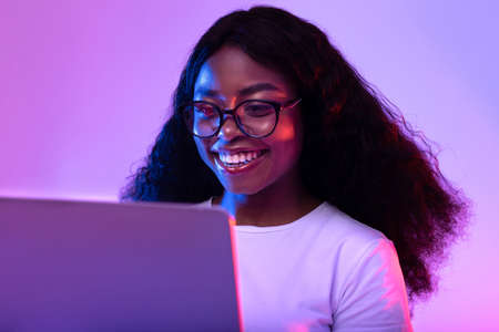 Happy young black woman in glasses using laptop pc for online work or studies in neon lightの写真素材