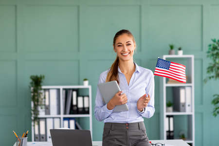 Happy foreign language teacher with USA flag standing near workdesk and holding digital tablet, smiling at cameraの写真素材
