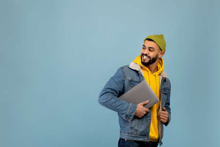 Online offer. Stylish arab man holding laptop and looking at copy space, hipster guy posing over blue backgroundの写真素材