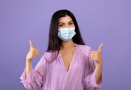 Portrait of armenian lady wearing medical face mask and gesturing thumbs up, posing to camera over...
