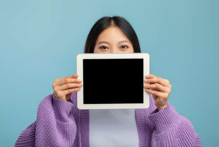 Young asian lady hiding her face behind digital tablet with empty black screen, showing cool mobile applicationの写真素材