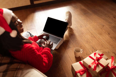 Black lady in Santa hat using laptop with empty screen, offering free space for holiday mockup advertisement at homeの写真素材