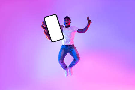 Full length of excited African American guy jumping with smartphone, demonstrating empty screen in neon light, mockupの写真素材